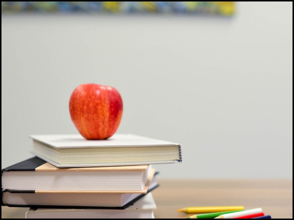 A stack of books on a classroom desk with a red apple on top